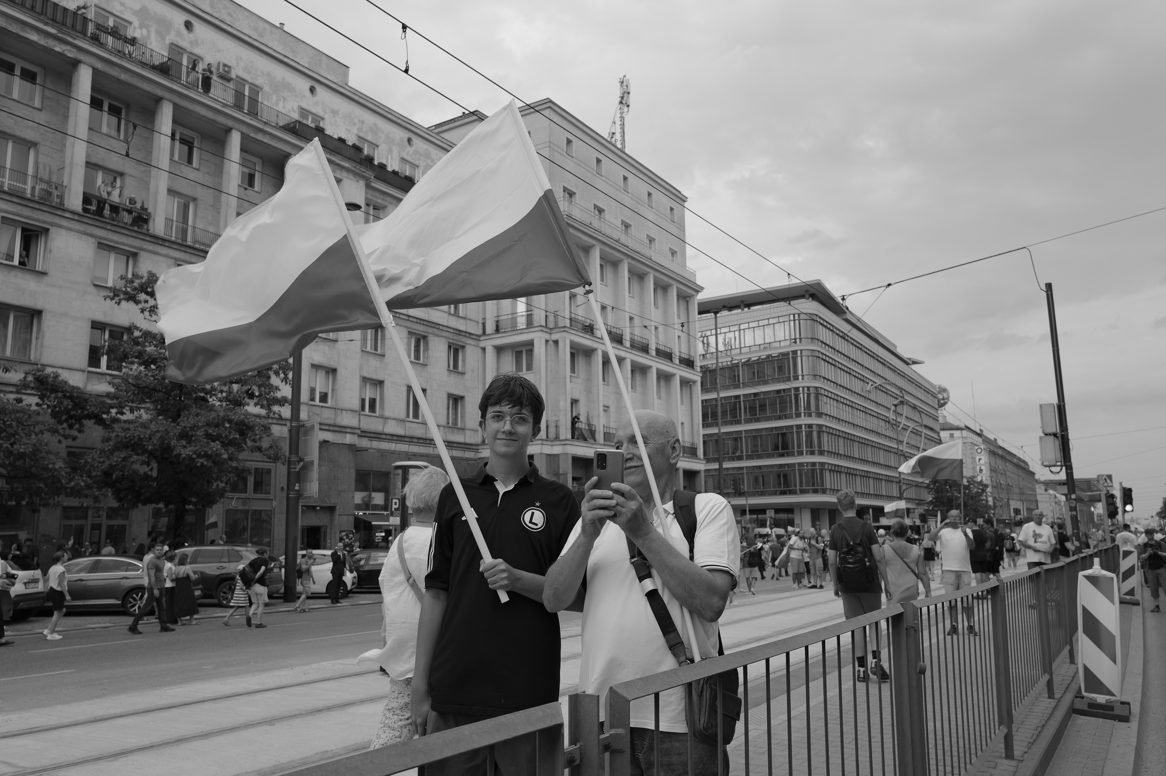 fot. Maciej Niedzielski, Czarno biala fotografia uliczna przedstawiająca dwóch mężczyzn trzymających polską flagę podczas godziny W w Warszawie wykonana Leica Q3